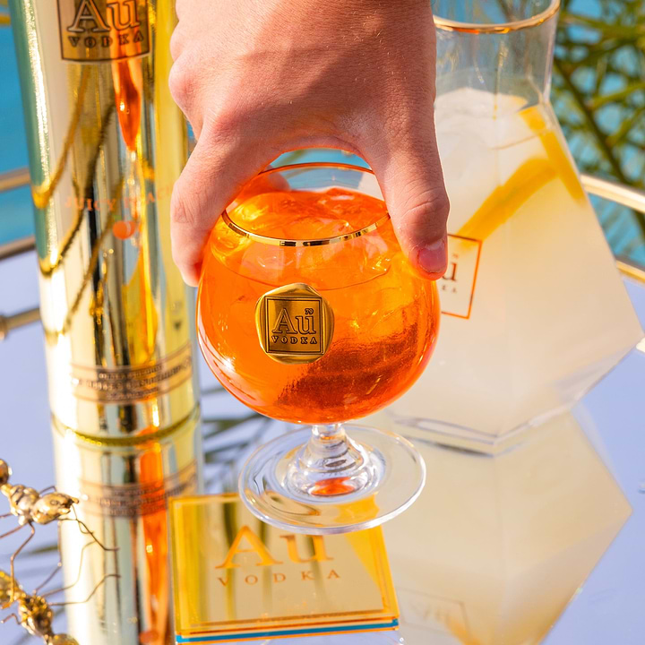 Hand holding an Au Vodka branded glass filled with orange cocktail and ice next to a gold bottle and carafe