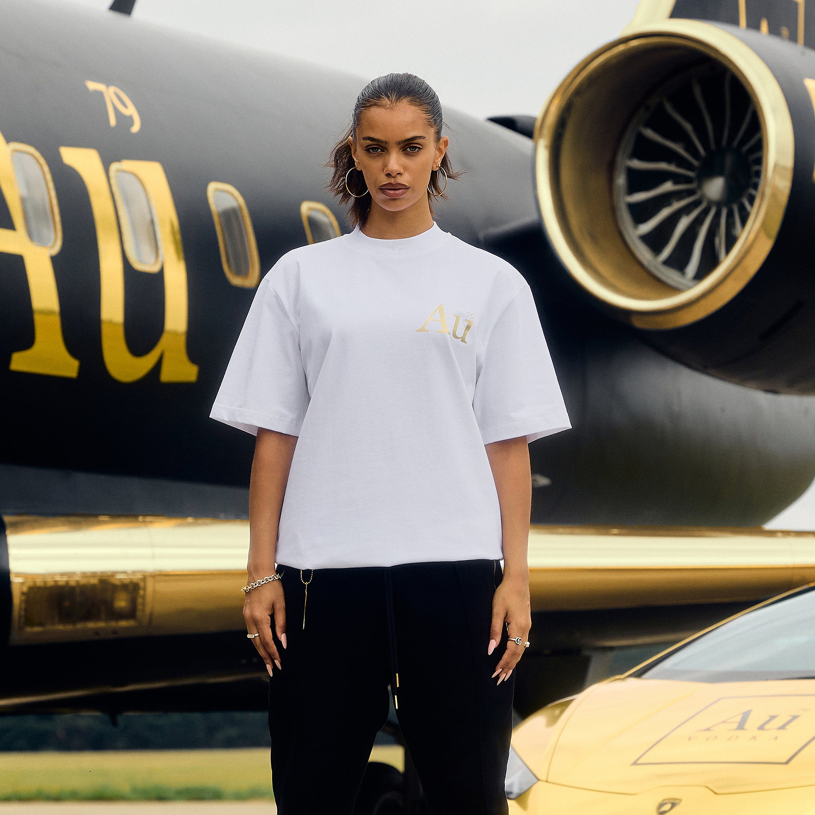 Model wearing white Au Vodka apparel t-shirt with gold logo in front of black private jet