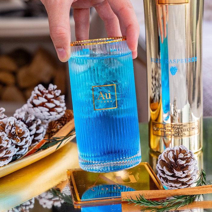 Hand holding a ribbed glass of blue Au Vodka Blue Raspberry cocktail next to a gold bottle and pinecones