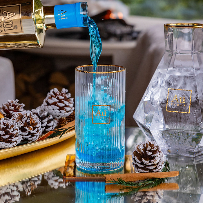 Au Vodka blue liquid pouring into a ribbed glass with ice on a table with pinecones and cinnamon