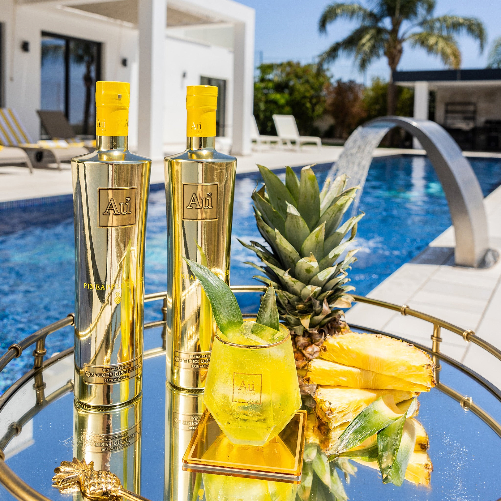 Two Au Vodka Pineapple Crush bottles, a cocktail, and pineapple slices on a mirrored tray by a pool