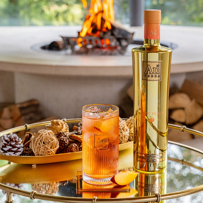 Au Vodka Icy Peach bottle next to a glass of peach cocktail on a gold tray with a fireplace in the background