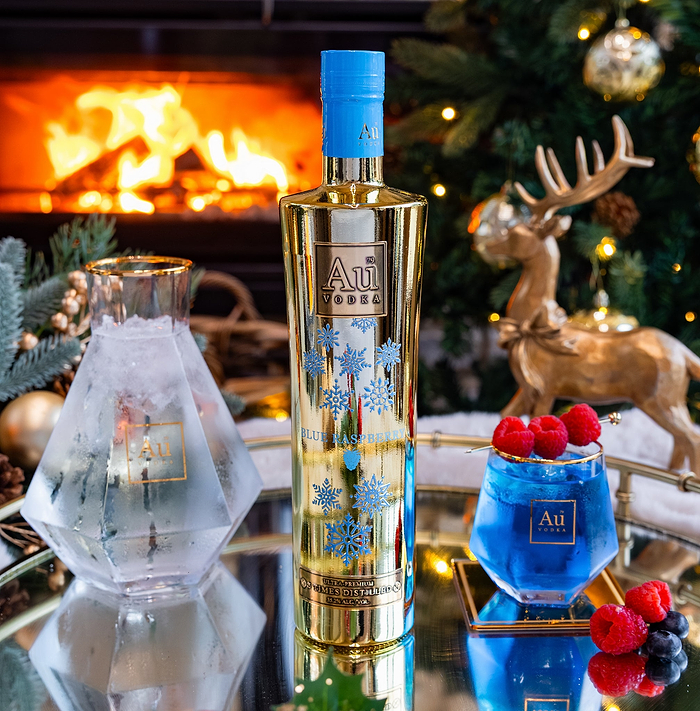 Christmas edition bottle of Au Vodka with decorative elements on a festive table with a fireplace and Christmas tree in the background.