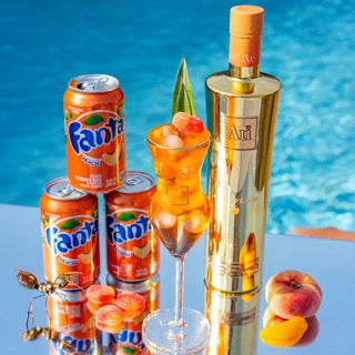 Au Vodka bottle with Fanta Peach cans and a cocktail glass on a reflective surface