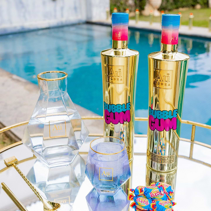 Two Bottles of Au Vodka Bubblegum, next to a clear jar of bubblegum sweets, and a Diamond Cut Glass, with bubblegum liquid, on a reflective surface. 