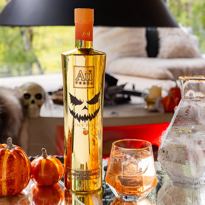 Gold bottle of Au Vodka with a jack-o'-lantern design on a table with pumpkins and a glass.