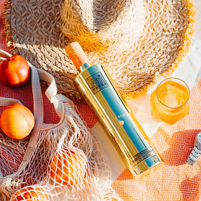 A Bottle of Au Vodka Juicy Peach, sitting on a beach towel, in a summery setting, next to a Au Diamond Cut Glass and other beach items like a Hat, A knitted bag. 