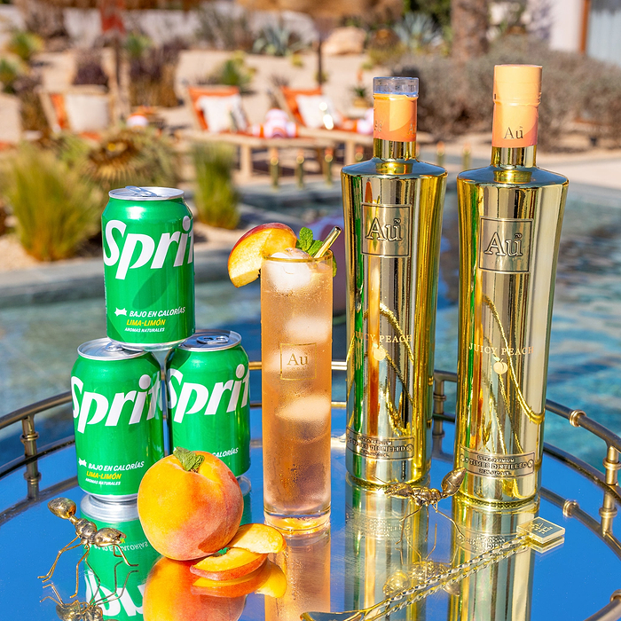 Two Bottles of Au Vodka Juicy Peach, next to an Au Highrise Glass filled with ice & orange colored liquid. Three Cans of Sprite, and a fresh peach, on a reflective surface. 