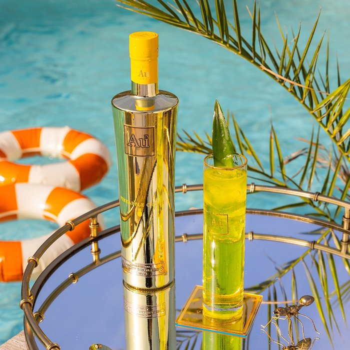 Au Vodka USA Pineapple Crush bottle and glass on a mirrored tray by a pool with a palm leaf