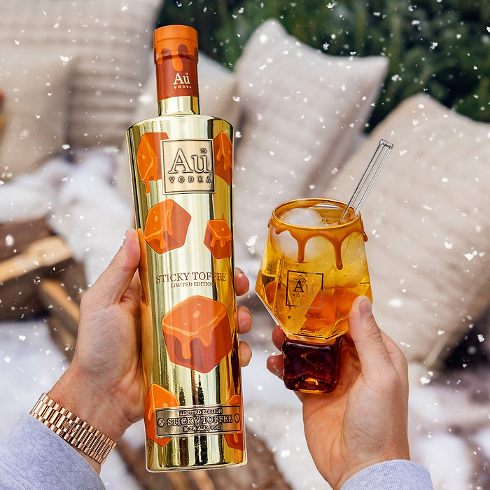 A Person Holding a Bottle of Au Vodka Sticky Toffee in one hand, and a Au Toffee Cube Glass in the other, against a blurred background.