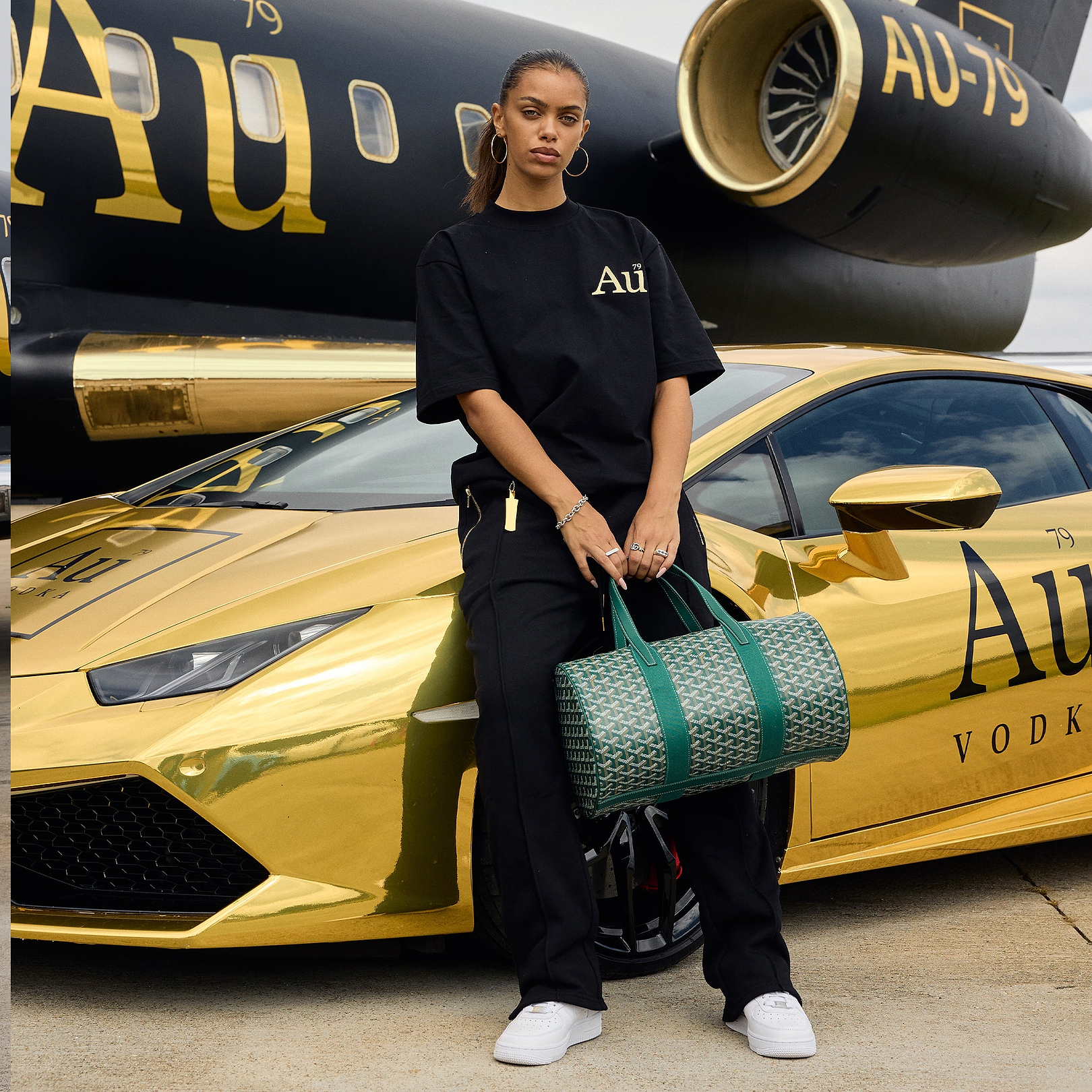 Person wearing black Au Vodka apparel standing in front of a gold sports car and private jet