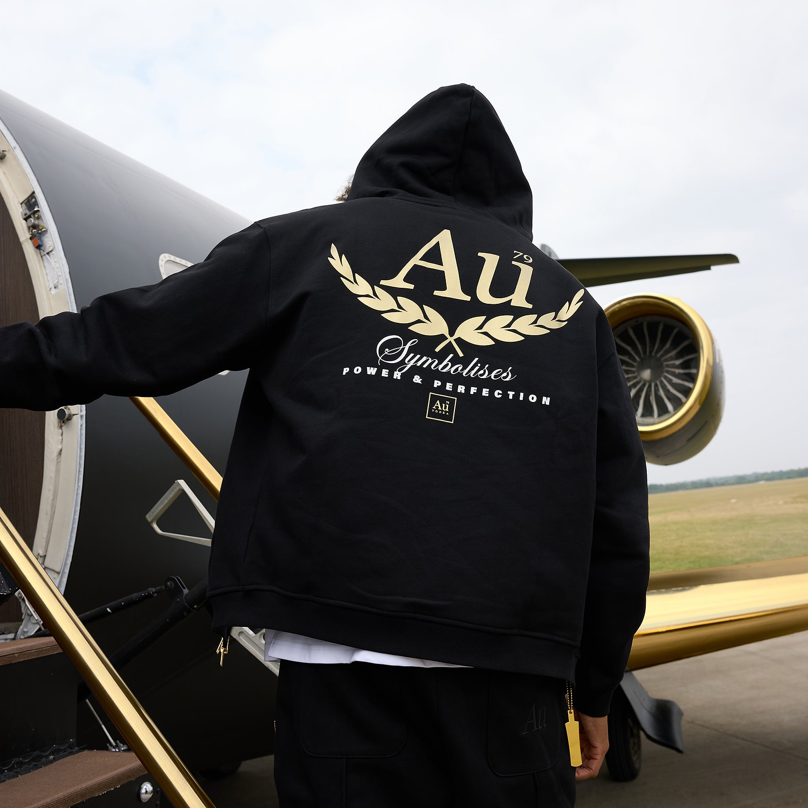 Person wearing black Au Vodka apparel hoodie with gold logo standing by private jet