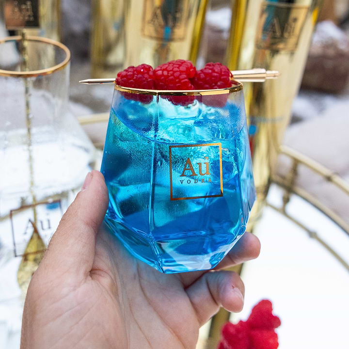 Hand holding a blue Au Vodka cocktail in a faceted glass garnished with raspberries