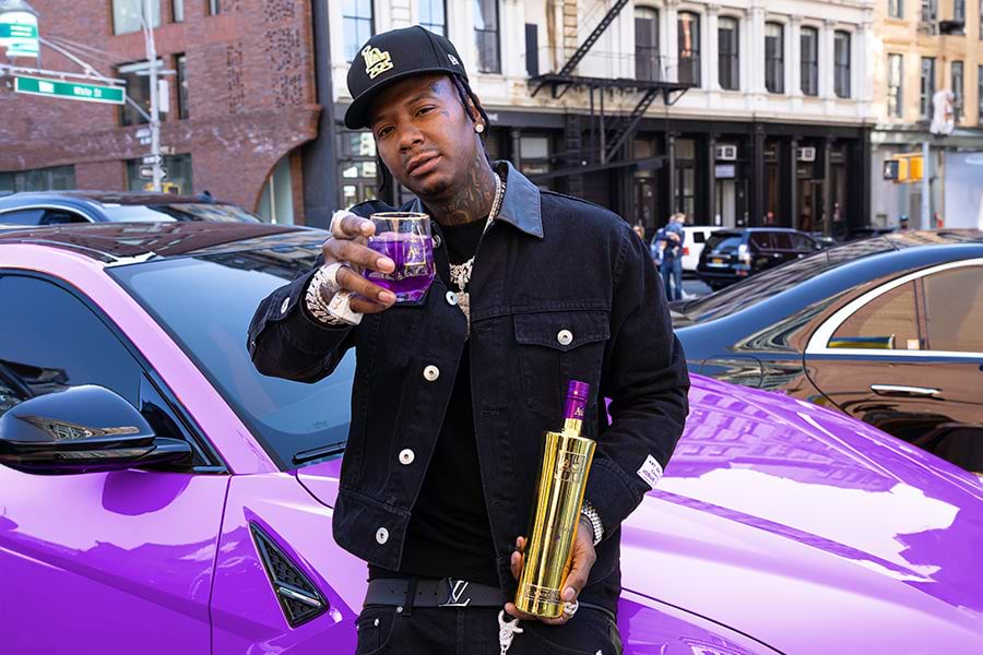 Man in black jacket and cap holding a purple drink and a gold bottle next to a purple car