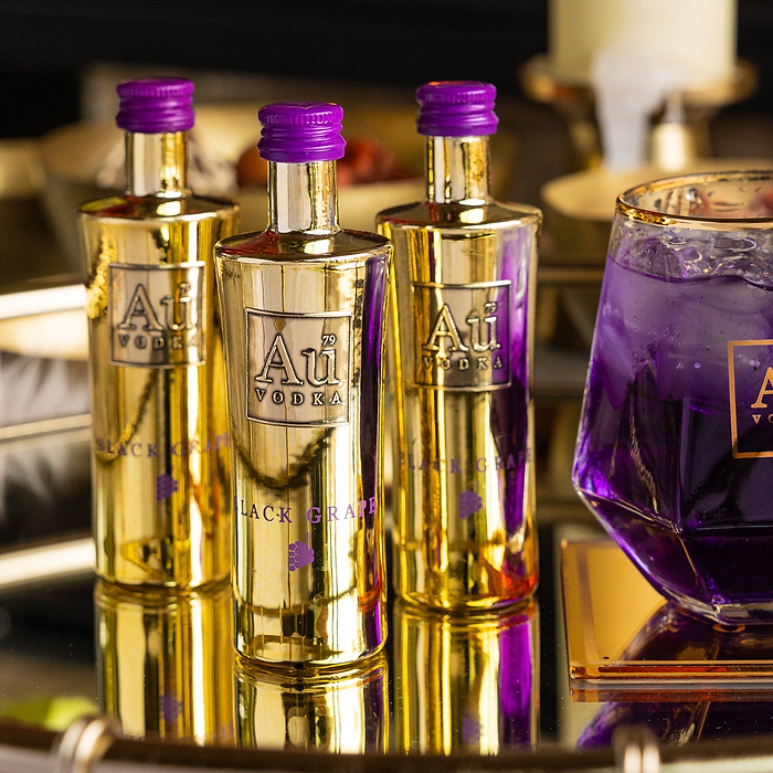 Three gold bottles of Au Vodka Black Grape with purple caps and a glass of purple cocktail on a reflective surface
