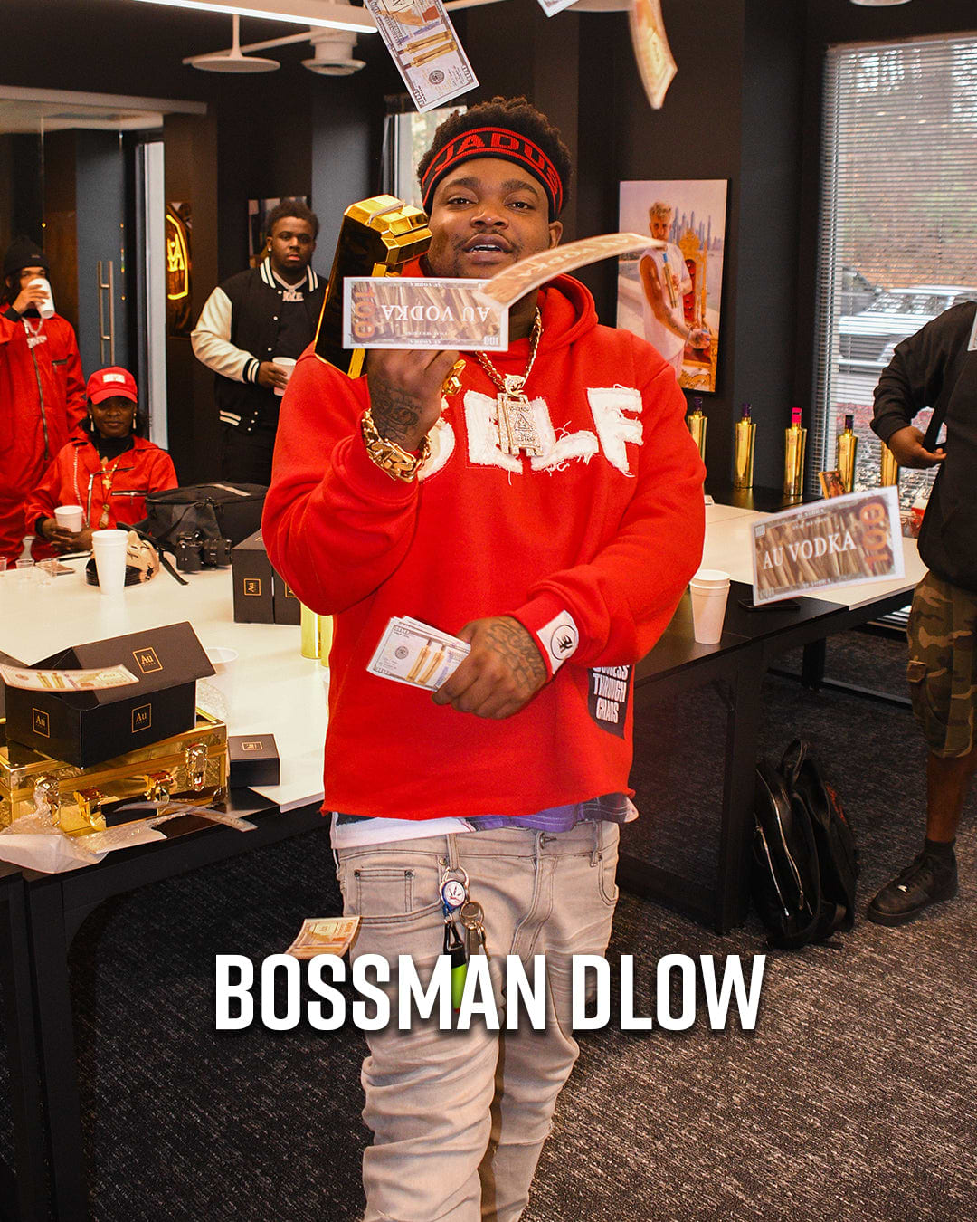 Bossman Dlow in a red hoodie holding money, with Au Vodka bottles on a table