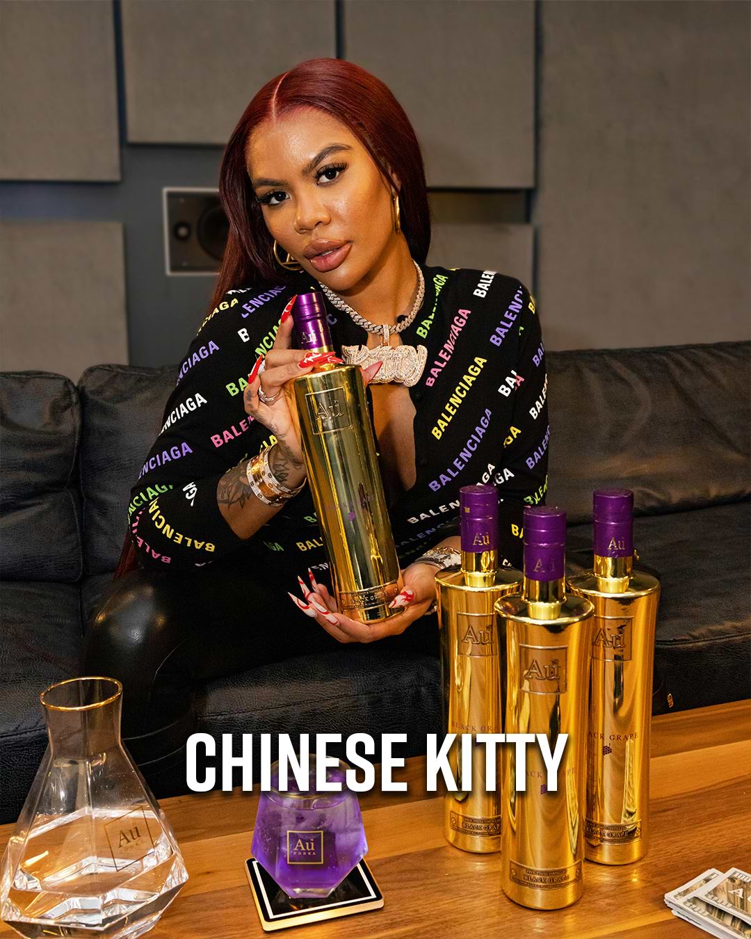 Chinese Kitty holding a gold Au Vodka bottle with purple accents, with other bottles on a table