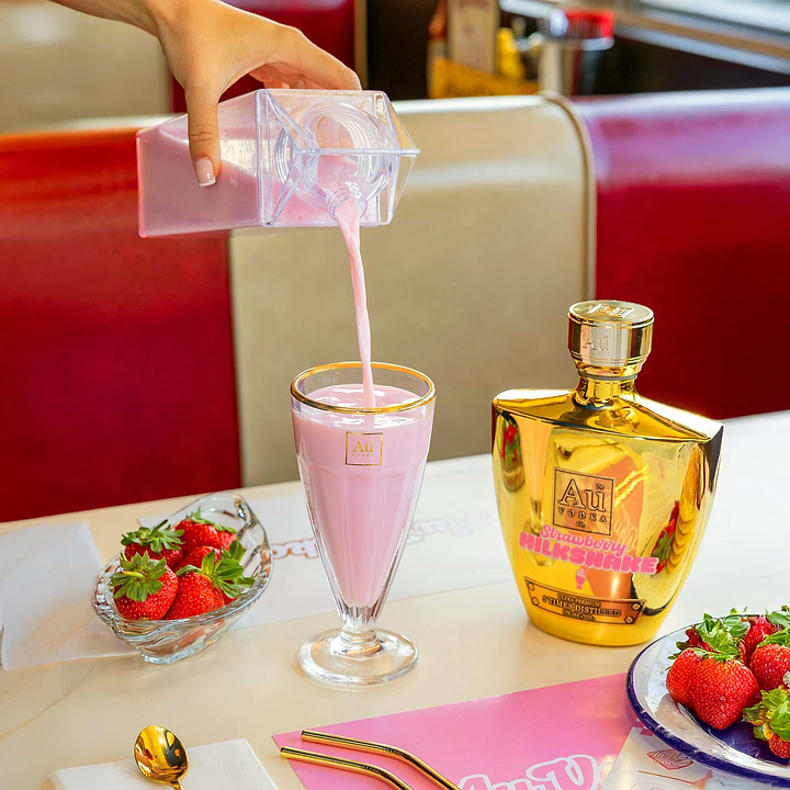 Hand pouring pink cocktail from a clear carton into a glass next to Au Vodka bottle