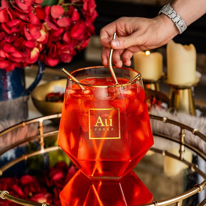 Au Vodka branded faceted glassware containing a red cocktail with strawberries and gold straws on a mirrored tray