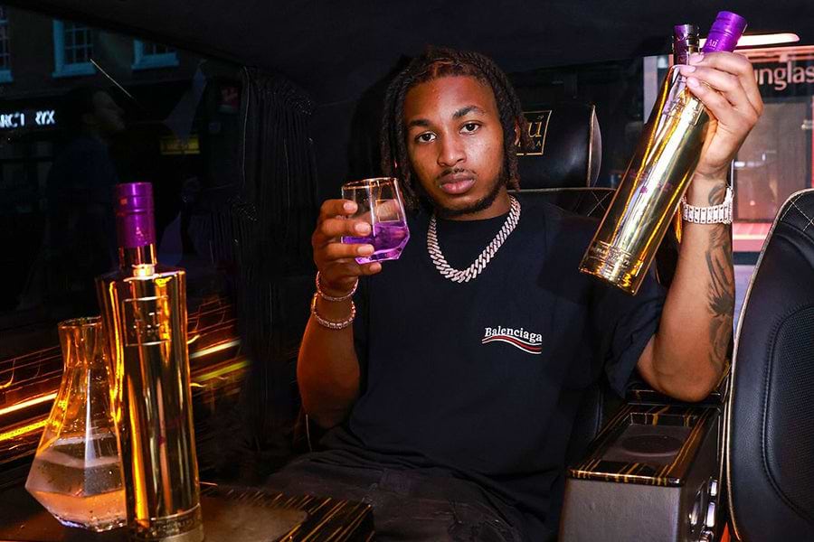 Man in a Balenciaga shirt holding a gold bottle and a glass with purple liquid