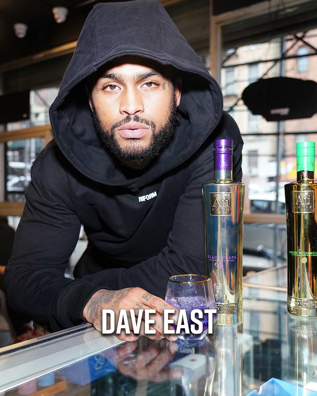 Dave East in a black hoodie with Au Vodka bottles and a drink
