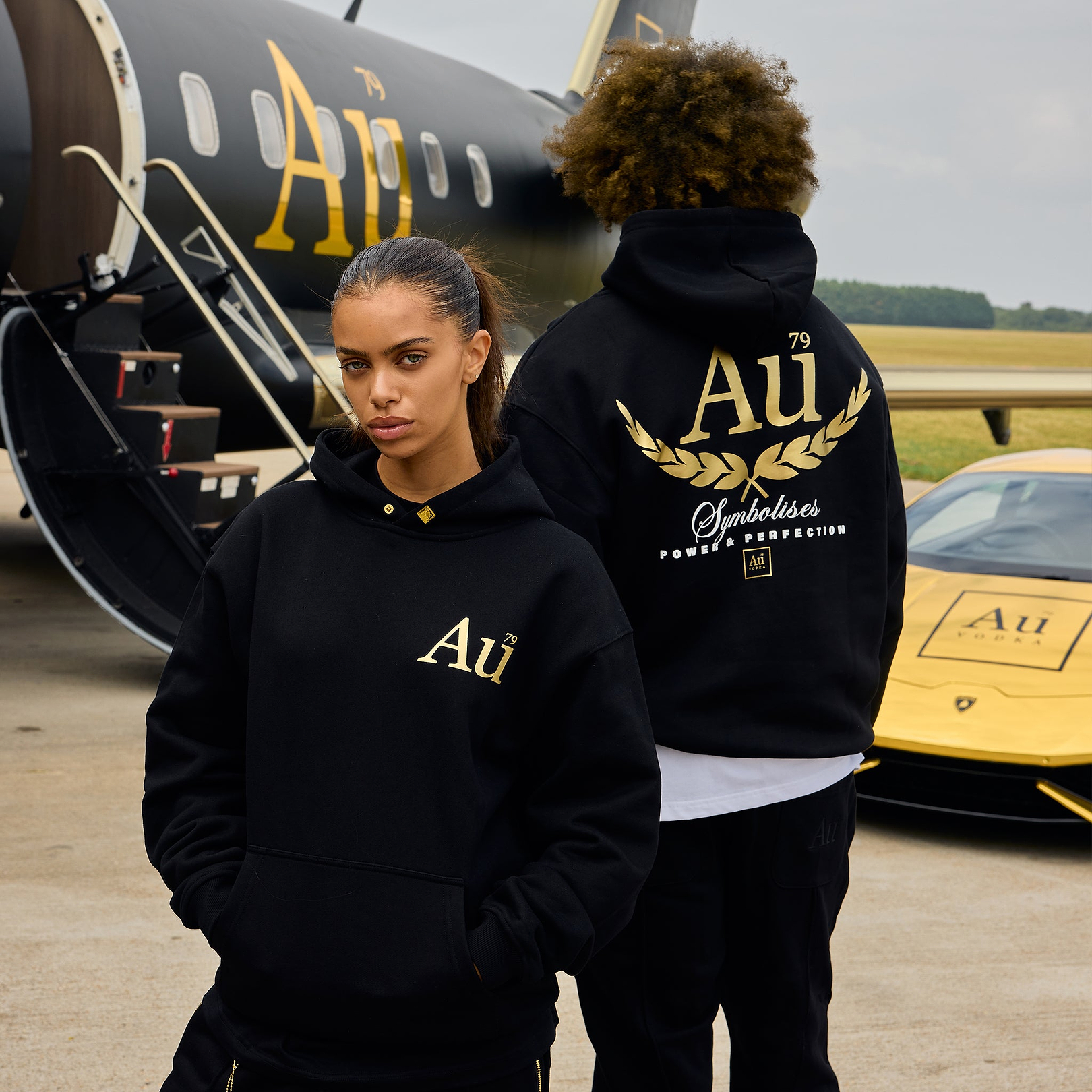 Two models wearing black Au Vodka branded hoodies standing in front of a private jet and a yellow sports car