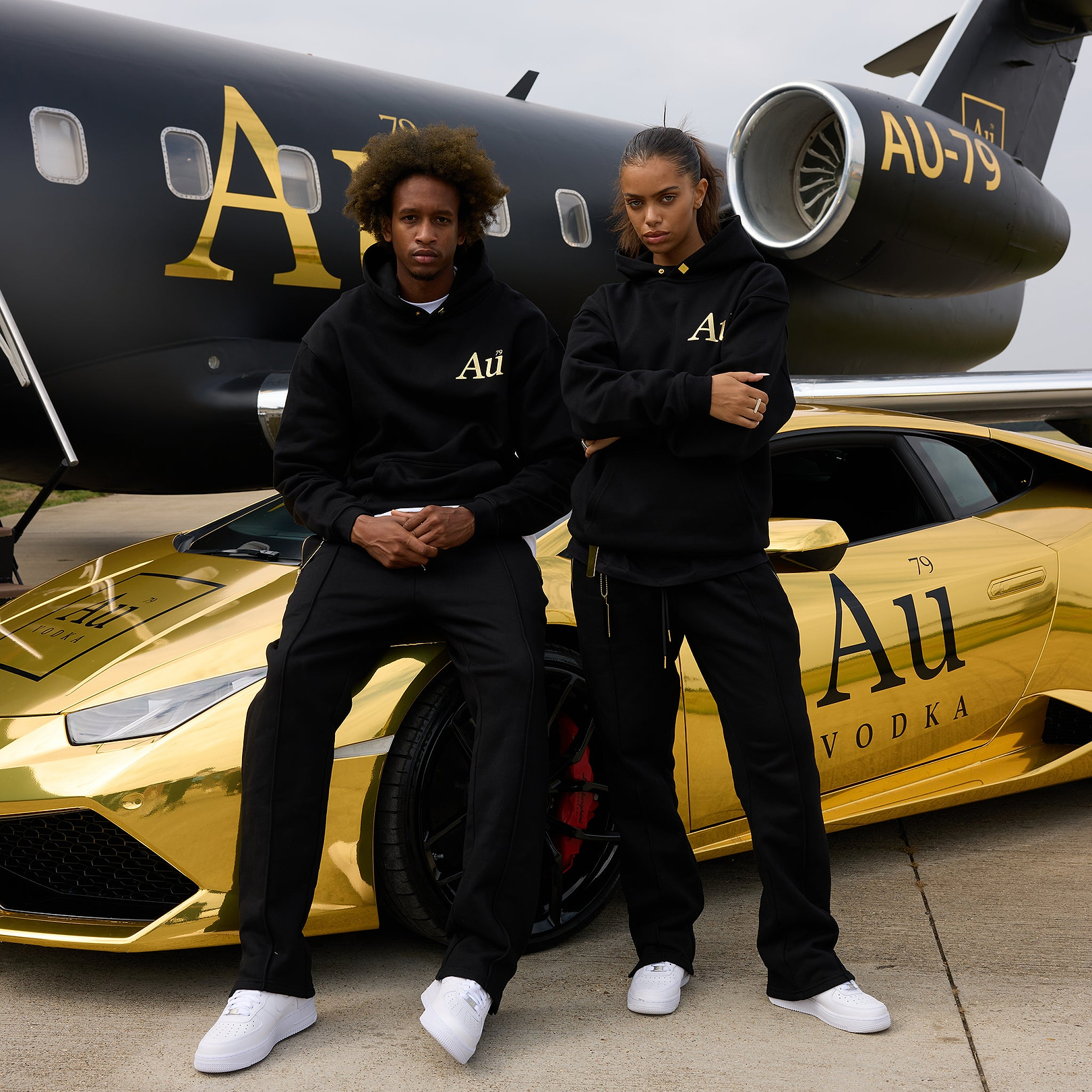 Two models wearing black Au Vodka branded hoodies and sweatpants standing by a gold car and private jet