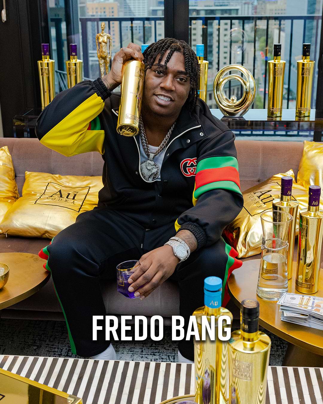 Fredo Bang holding a gold Au Vodka bottle, surrounded by more bottles and gold pillows