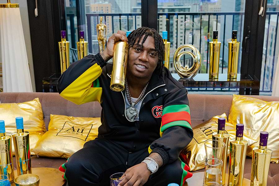 Man holding a gold Au Vodka bottle, surrounded by more bottles and gold pillows