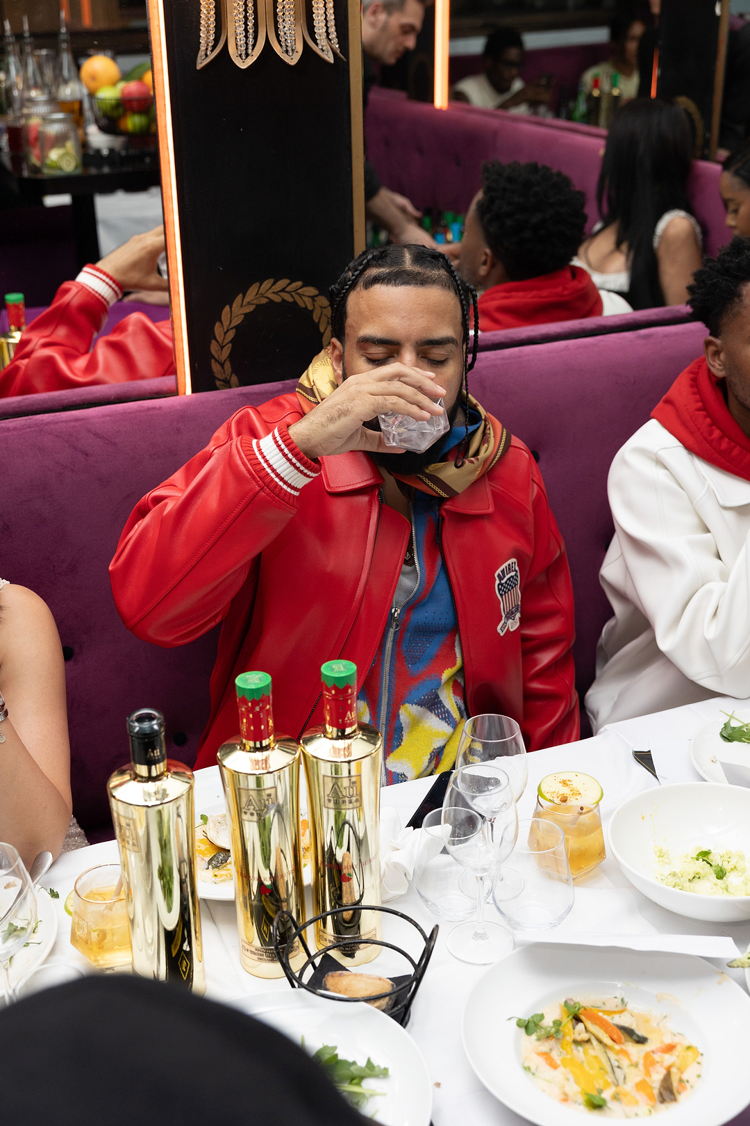 Man in red jacket drinking from a glass at a table with Au Vodka bottles and food
