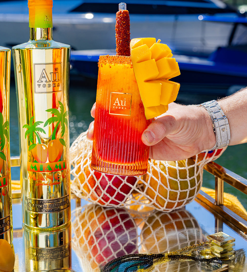 Hand holding a mango vodka cocktail with Au Vodka Miami Limited Edition bottle in the background