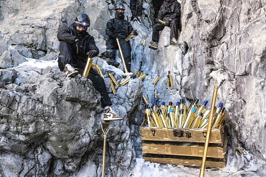 Three people in black gear and headlamps on a rocky, snowy cliffside with gold bottles and a gold trunk