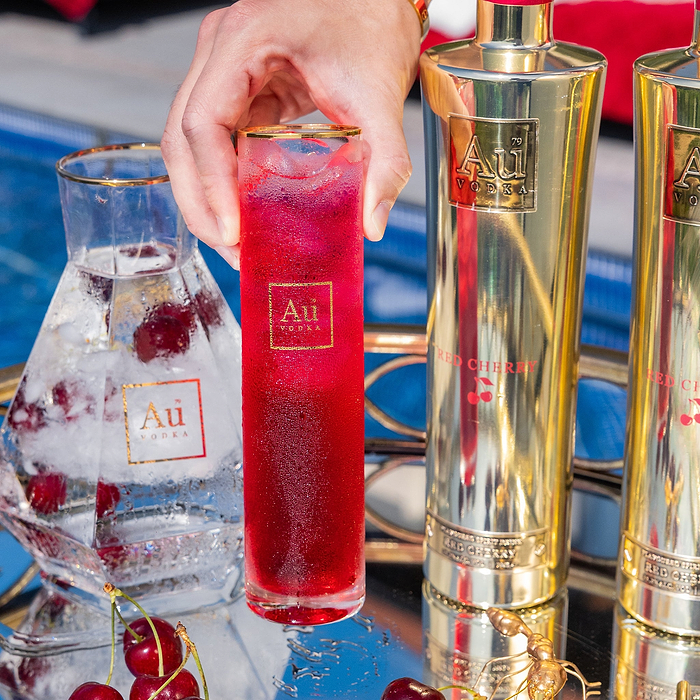 Hand holding an Au Vodka branded highball glass filled with a red cherry cocktail next to Au Vodka bottles