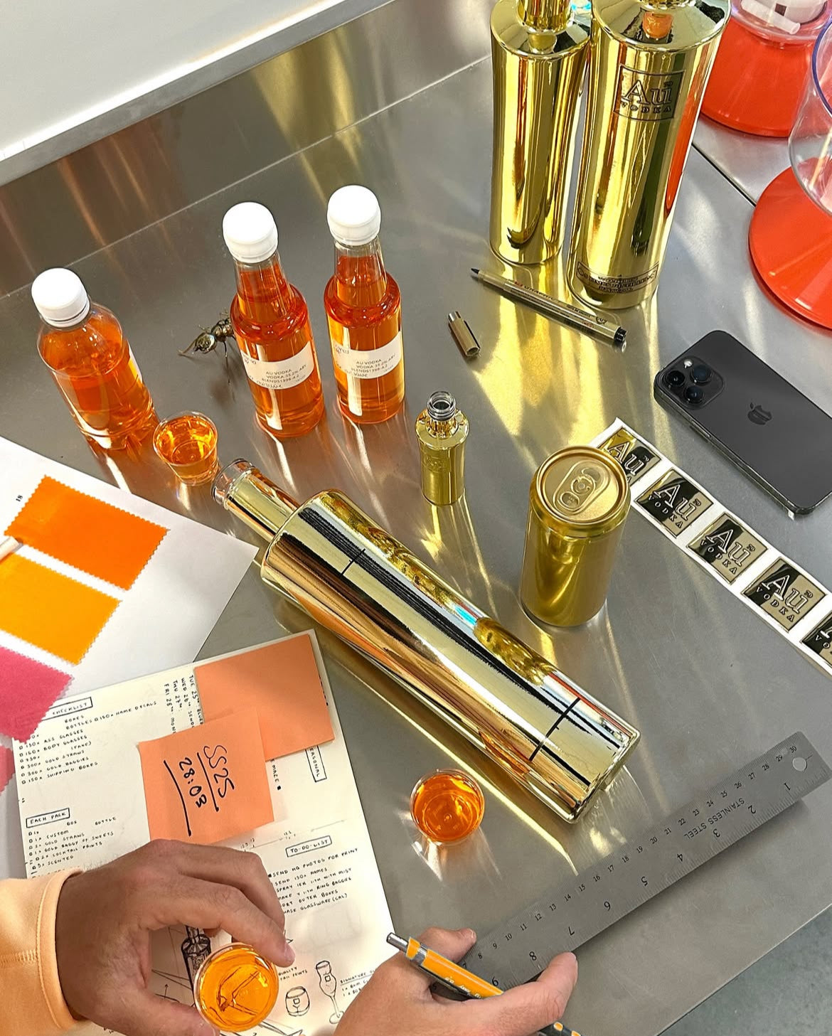Au Vodka gold bottles and cans with orange liquid samples on a metal workspace with a checklist and ruler