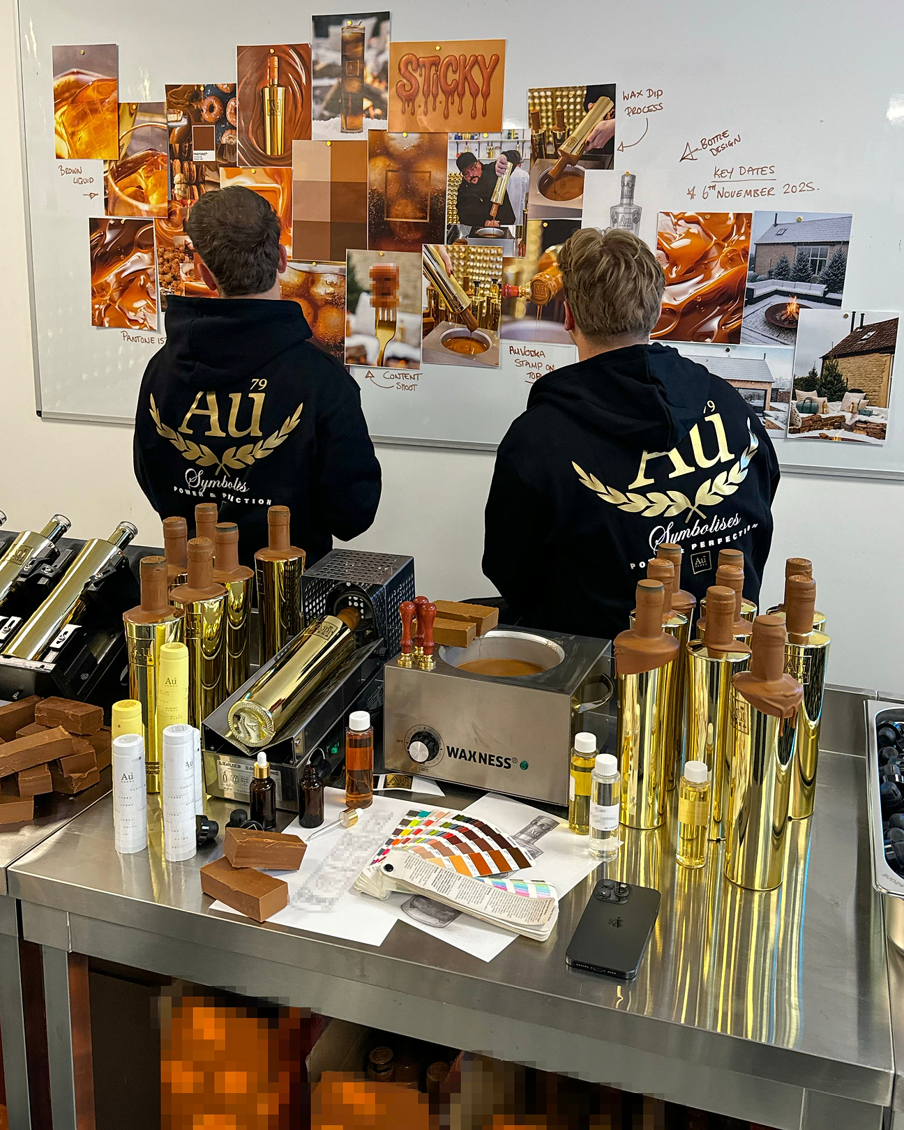 Two people wearing Au Vodka hoodies standing before a mood board with gold bottles and wax dipping equipment on a table
