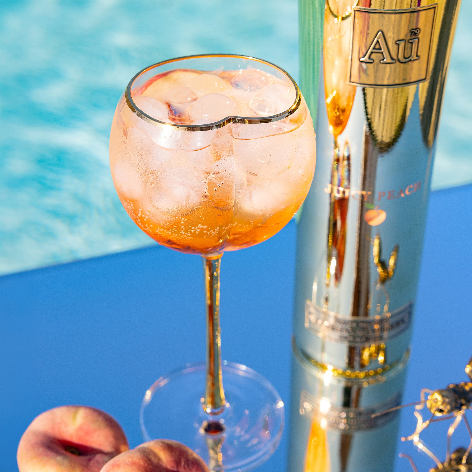 Au Vodka Juicy Peach cocktail in a gold rimmed glass next to a bottle and fresh peaches by a pool