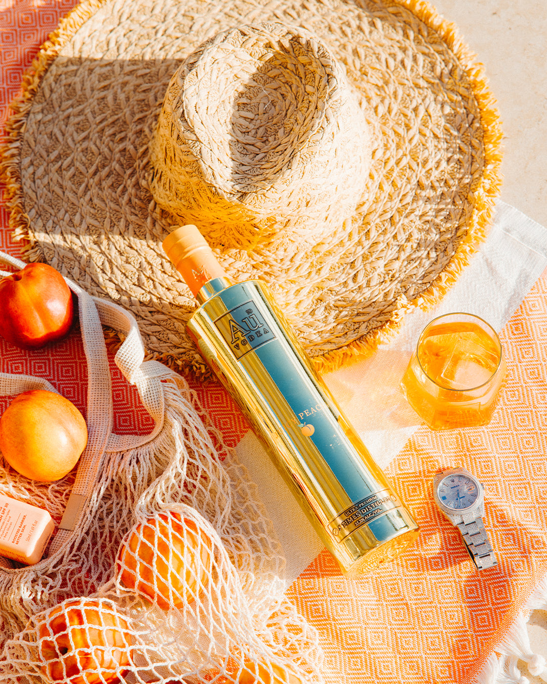 Au Vodka Peach bottle with fresh peaches, straw hat, and a glass of drink on an orange patterned picnic blanket