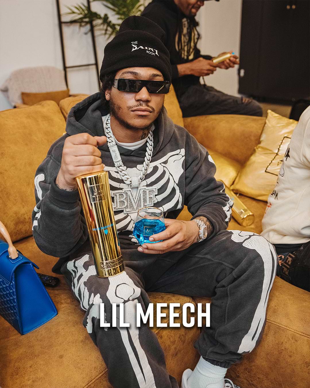 Lil Meech in a skeleton hoodie and beanie, holding a gold bottle and a glass with blue liquid