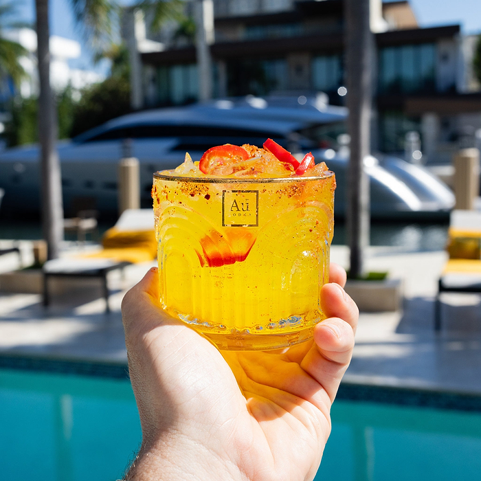 Hand holding a yellow Au Vodka cocktail with red chili garnish by a pool and yacht