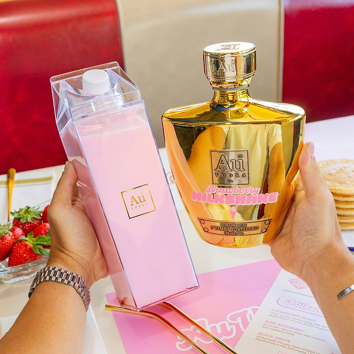 Hands holding a clear carton of pink cocktail and a gold Au Vodka bottle