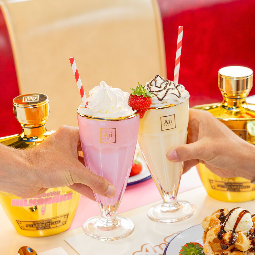 Two hands toasting with Au Vodka Strawberry Milkshake cocktails in tall glasses with gold bottles in the background