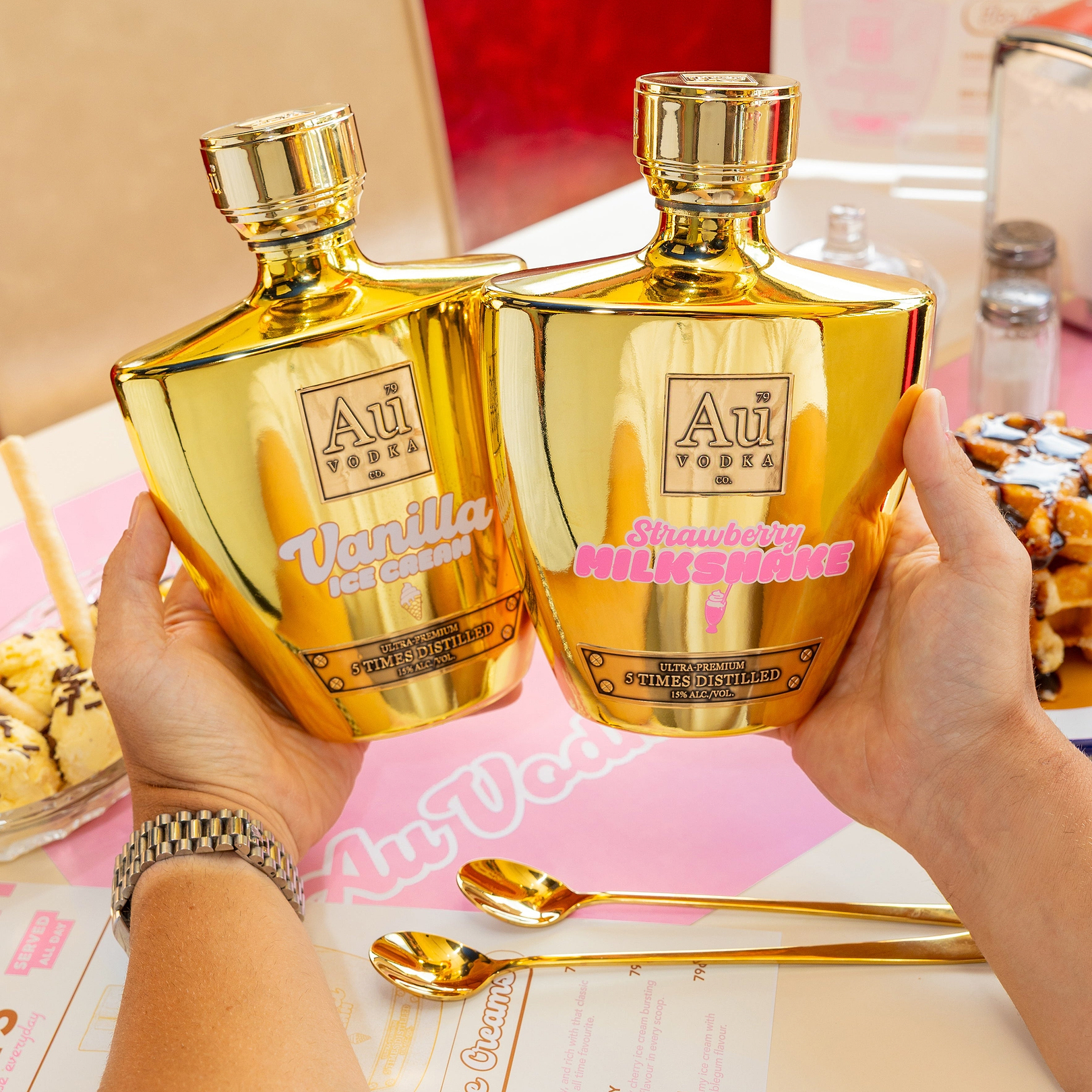 Hands holding Au Vodka Vanilla Ice Cream and Strawberry Milkshake bottles on a table with dessert