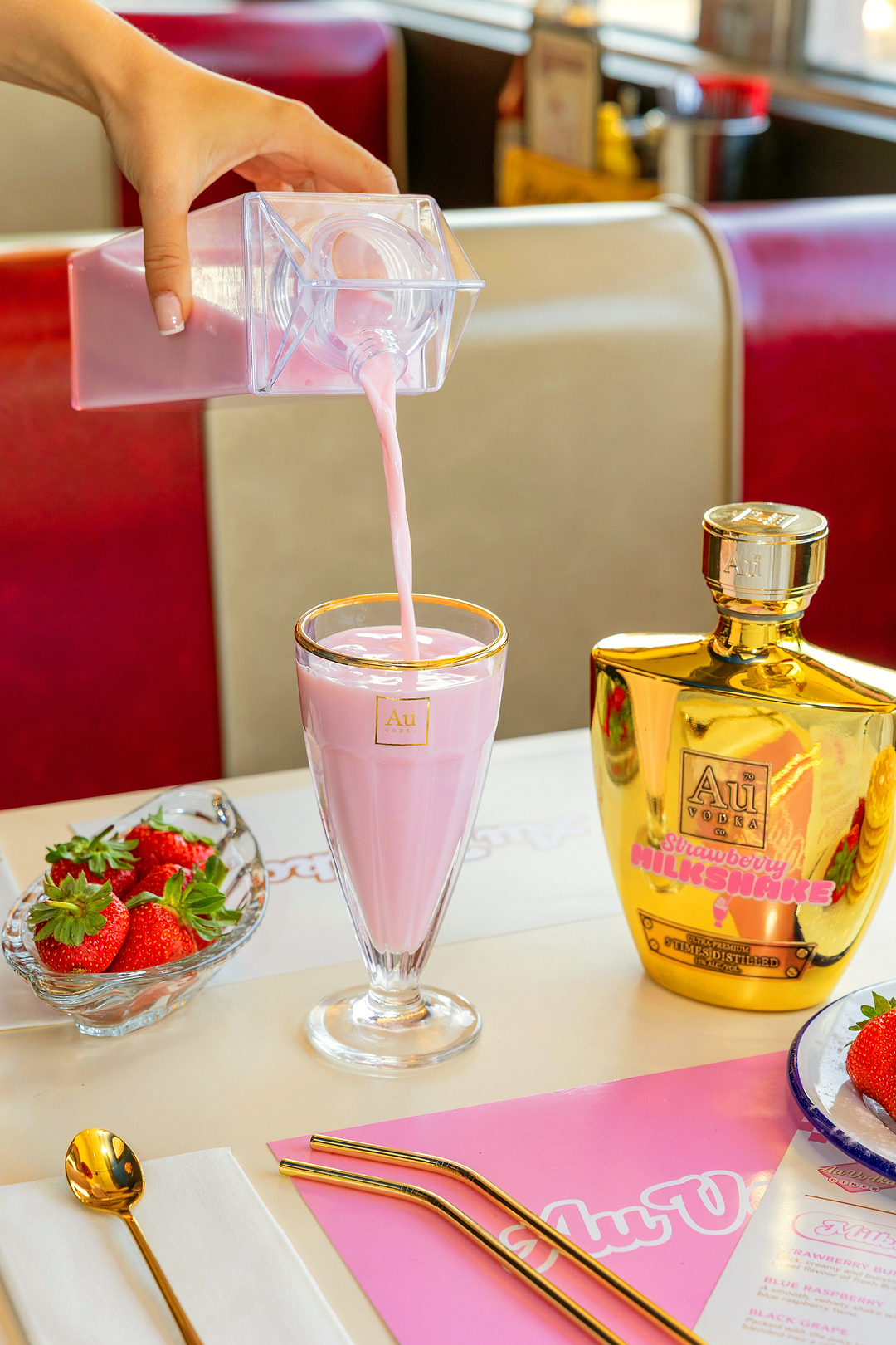 Hand pouring pink Au Vodka Strawberry Milkshake into a glass next to a gold bottle and fresh strawberries