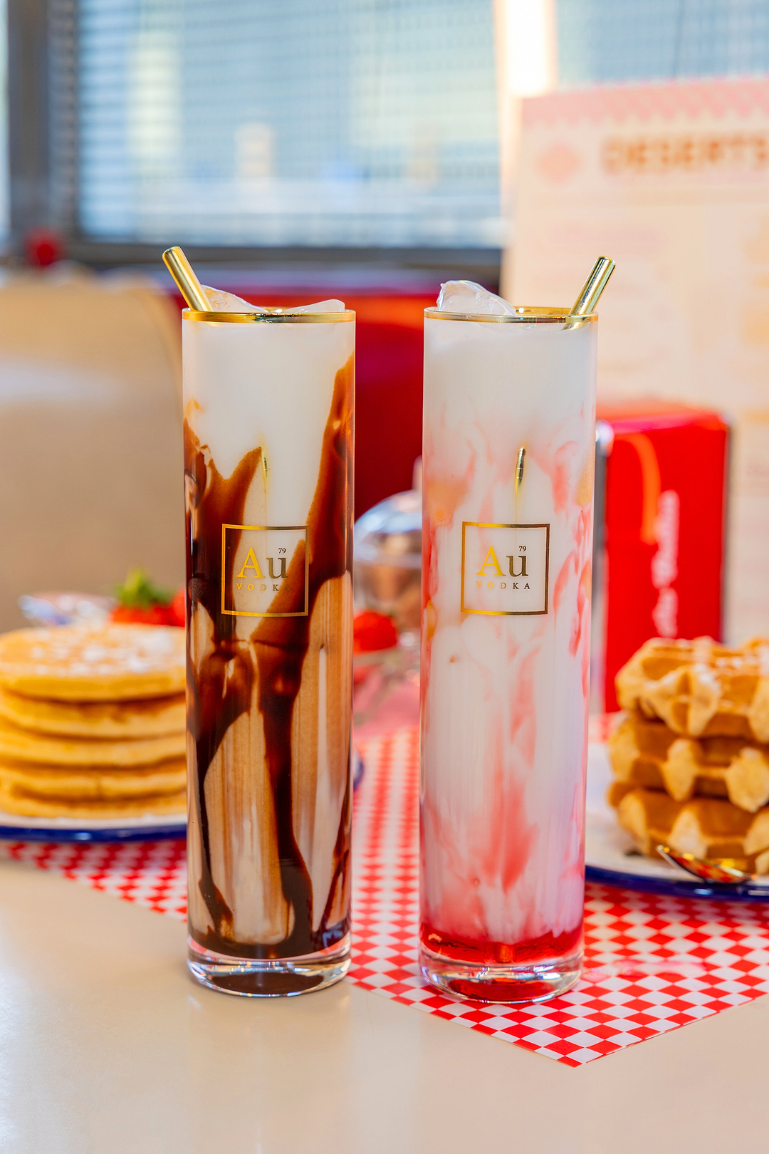Two tall Au Vodka glasses with chocolate and strawberry milk drinks on a checkered placemat with waffles