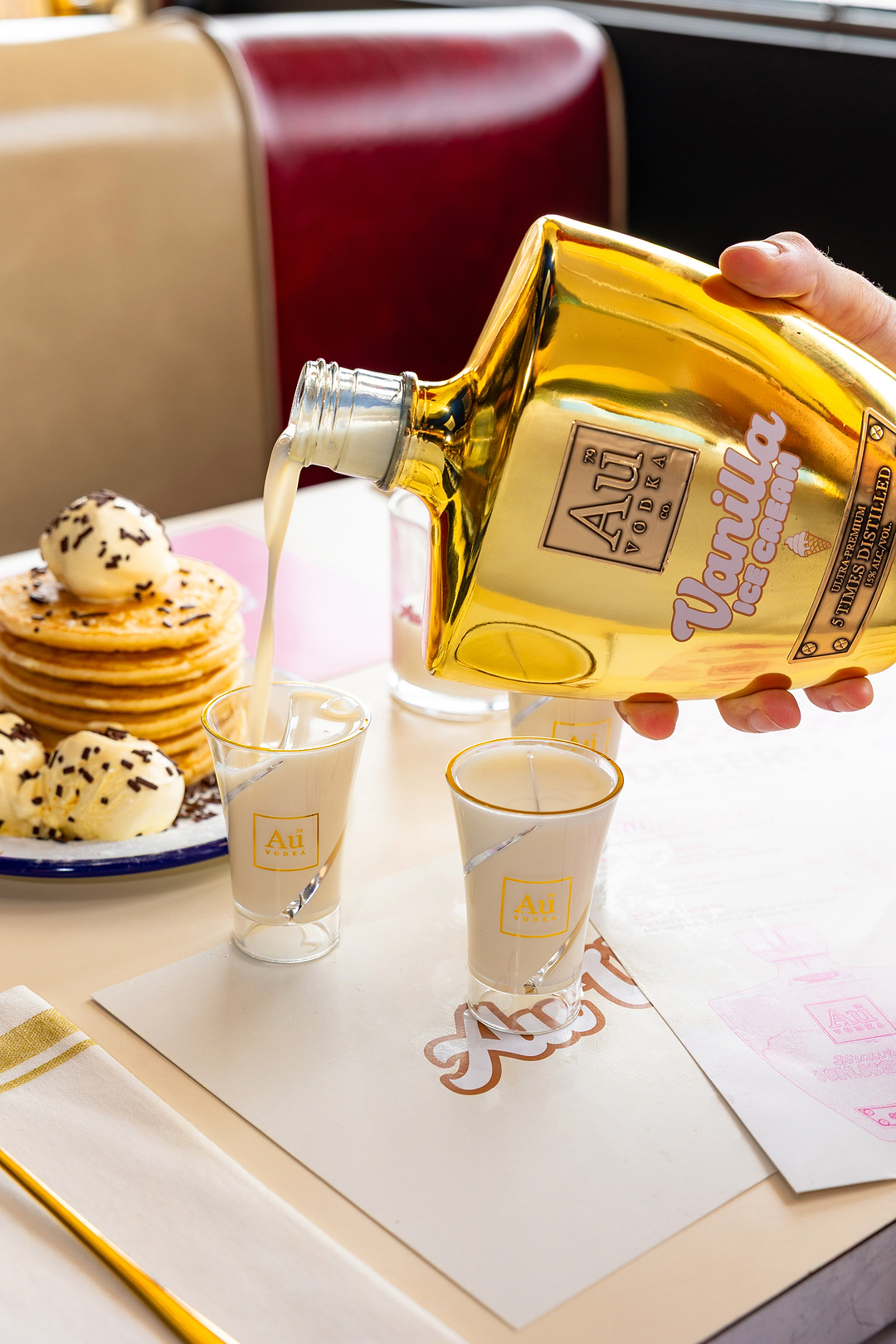 Au Vodka Vanilla Ice Cream being poured into shot glasses next to a stack of pancakes
