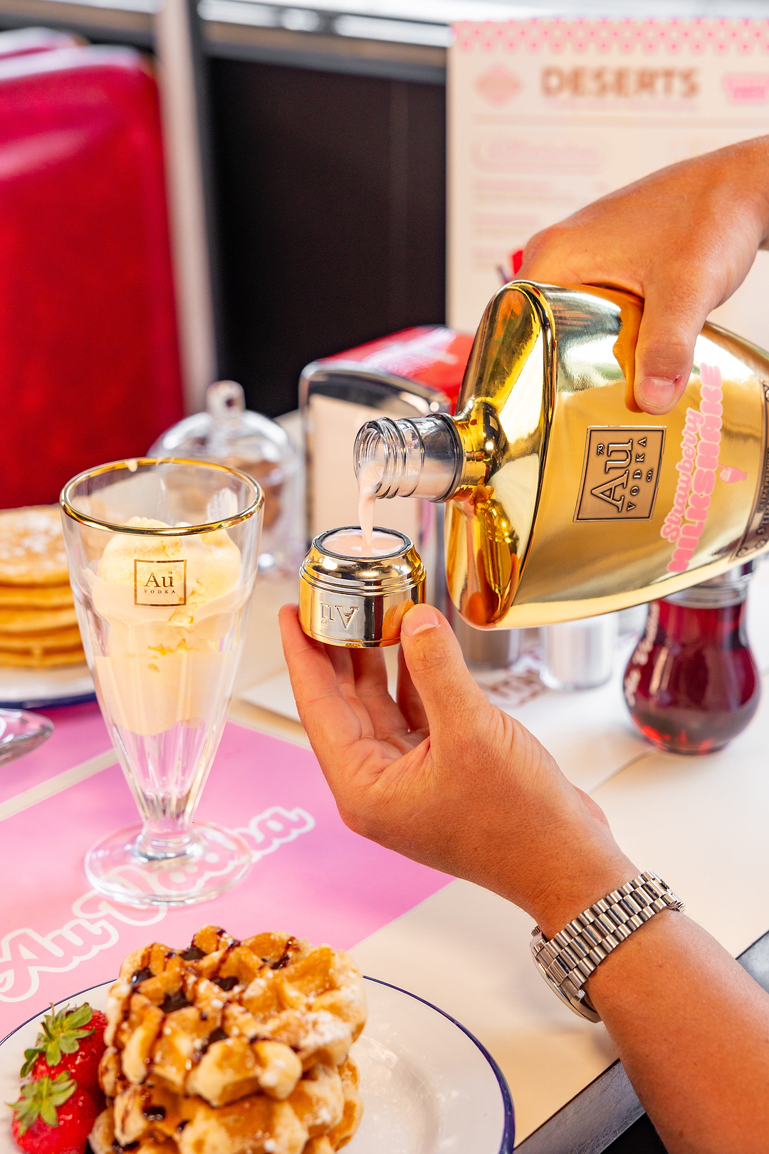 Au Vodka Strawberry Milkshake being poured into a gold shot glass at a diner table with waffles and ice cream