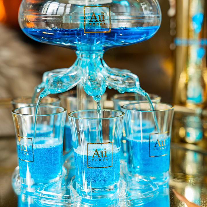 Close up of an Au Vodka glass shot tower filled with blue liquid on a mirrored tray