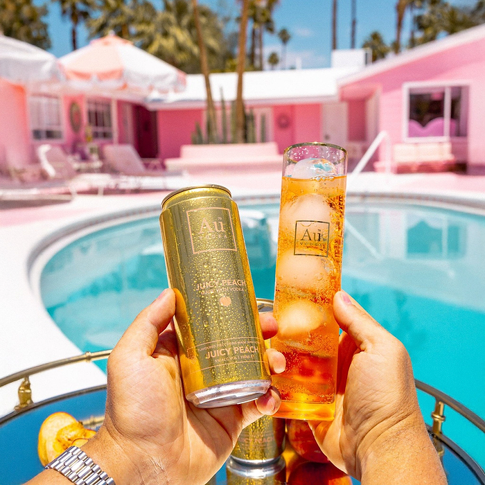 Hands holding an Au Vodka USA Juicy Peach can and a tall glass with ice and drink