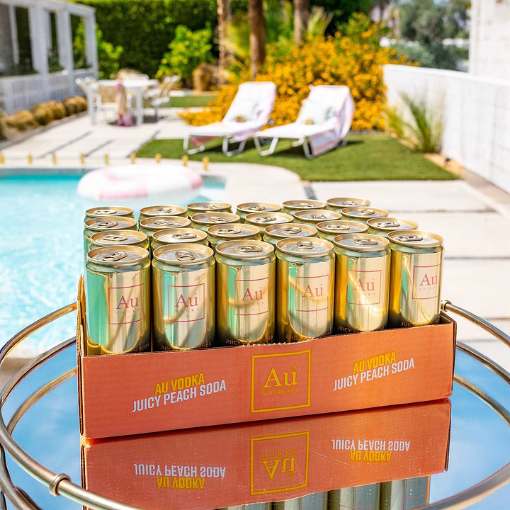 Case of Au Vodka Juicy Peach Soda cans on a mirrored table by a swimming pool