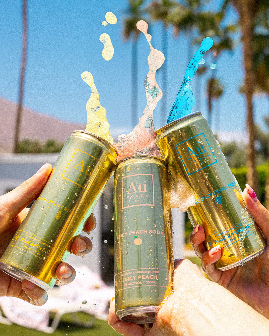 Three Au Vodka cans in Pineapple Crush, Juicy Peach, and Blue Raspberry splashing against a sunny outdoor background